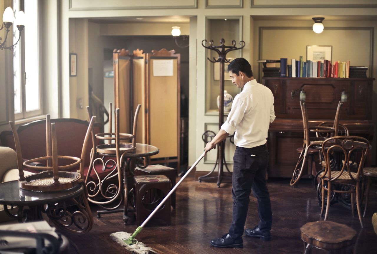 about-img Side view of young ethnic male in uniform cleaning floor with mop while working in retro styled restaurant