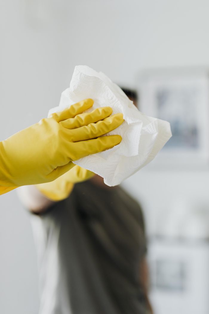 about-01 Homme anonyme portant des gants en latex jaunes et un t-shirt sombre, essuyant un miroir dans une salle de bain avec une serviette en papier